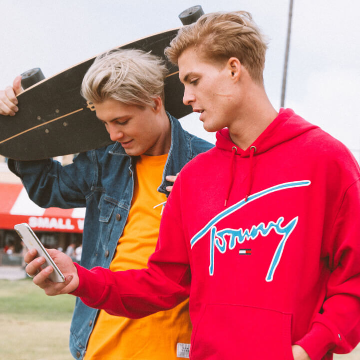 Two male models, one model wearing Tommy Hilfiger jacket and XPLORE t-shirt and other model wearing XPLORE sweatshirt holding a cell phone.